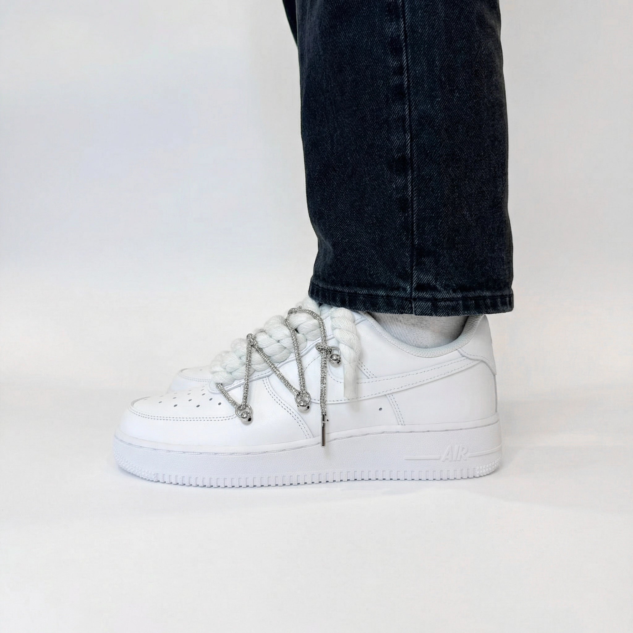 Bočný pohľad na biele Nike Air Force 1 s bielymi rope laces a striebornými ozdobami na nohách